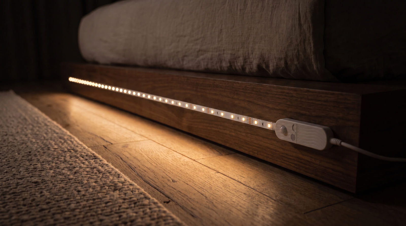 A light strip mounted under a bed frame, light pooling on an oak floor